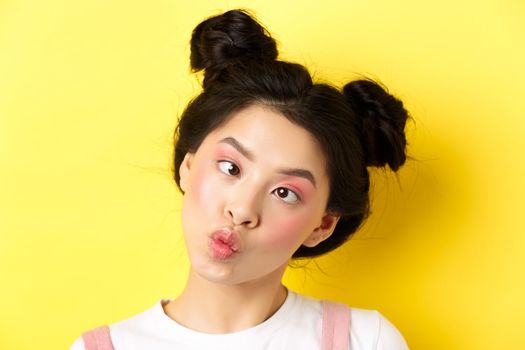 Close up portrait of funny asian teen girl with glamour makeup and hairstyle, squinting eyes and pucker lips silly, standing on yellow background.