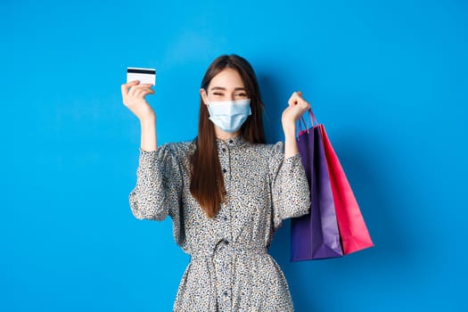 Covid-19, pandemic and lifestyle concept. Pretty female model in medical mask, rejoicing over shopping discounts, holding plastic credit card and bags, standing happy on blue background.