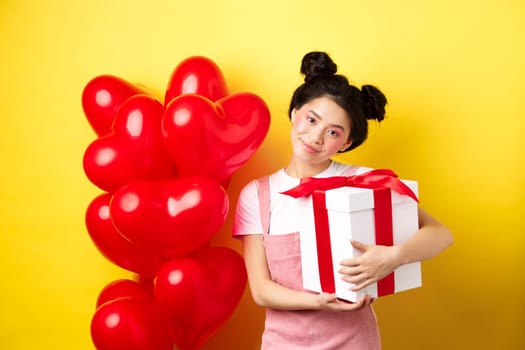Happy Valentines day. Cute asian girl hugging surprise gift from boyfriend, standing near lovely red heart balloons and yellow background.