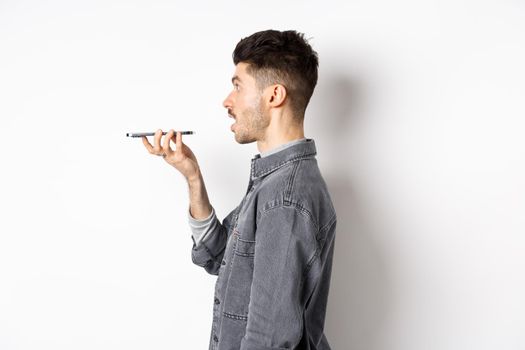 Profile of young man speaking at smartphone, translate with voice-translator app, talking into mobile phone speakerphone, standing on white background.