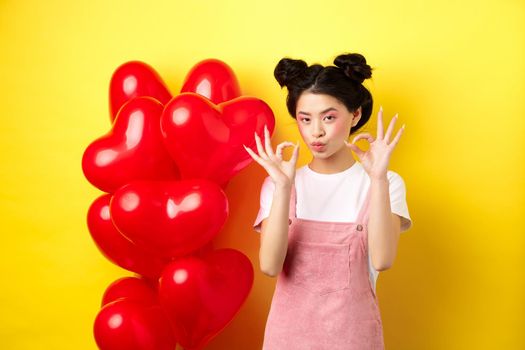 Cute young asian woman in stylish clothes and makeup, showing okay signs near st Valentines day heart balloons, praise good offer, standing on yellow background.