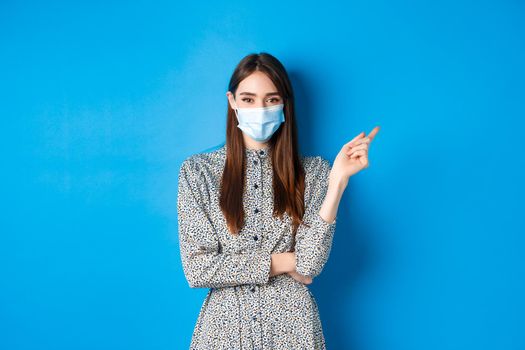 Covid-19, social distancing and healthcare concept. Pretty lady in medical mask showing way, pointing right and smiling, advertising on blue background.