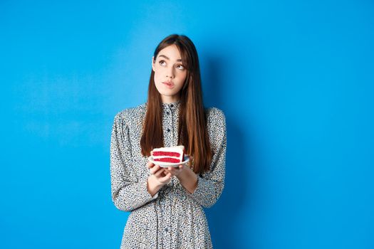 Birthday girl making wish, looking aside at logo and holding b-day cake, standing on blue background.