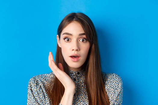 Candid girl with long hair and makeup, looking shocked and surprised, open mouth and stare speechless at camera, standing on blue background.