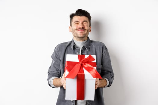 Happy handsome young man holding big valentines day gift and cheering, receive romantic surprise present from lover, standing on white background.