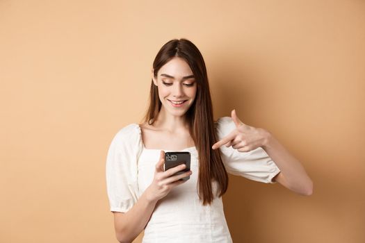 Smiling cute girl reading screen and pointing at mobile phone, standing on beige background.