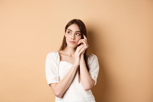 Dreamy and romantic girl looking aside with thoughtful gaze, touching clean gentle facial skin, standing against beige background.