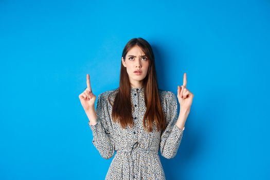 Confused attractive woman in dress frowning, pointing fingers up and looking puzzled, cant understand something, stare at strange thing, blue background.