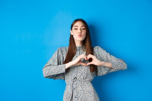 Valentines day. Romantic girl pucker lips for kiss, showing heart gesture, say I love you and look at camera, standing in dress on blue background.