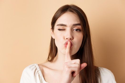 Close up of attractive woman shushing at camera, hiding secret, making hush sign and winking, show taboo sign, standing on beige background.