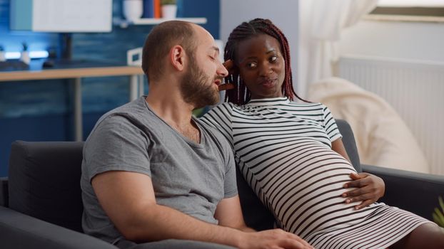 Interracial couple with pregnancy at home relaxing on living room sofa. Young caucasian father of child holding hand on belly while african american pregnant mother laying on couch