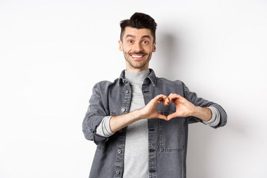 Smiling boyfriend showing heart with love, looking romantic and happy at lover, standing on white background. Valentines day and relationship concept.