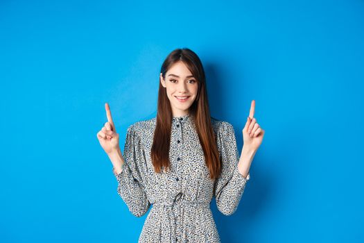 Cute young woman with long hair and vintage dress, smiling and pointing fingers up, advertising on blue background.