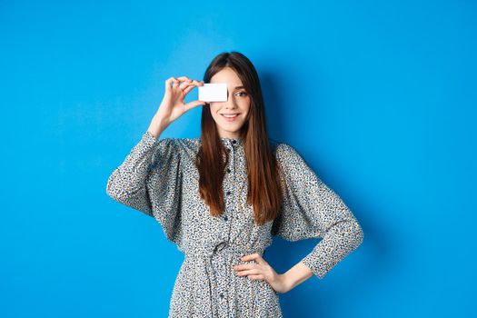 Shopping. Beautiful adult woman with natural long hair showing plastic credit card over eye, standing against blue background.