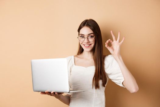 Freelance girl in glasses showing okay sign and working on laptop, guarantee good quality, recommend something online, standing on beige background.