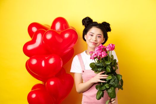 Happy Valentines day. Beautiful asian woman dressed for romantic date, holding bouquet of flowers and smiling. Girl with roses standing near heart balloons, yellow background.