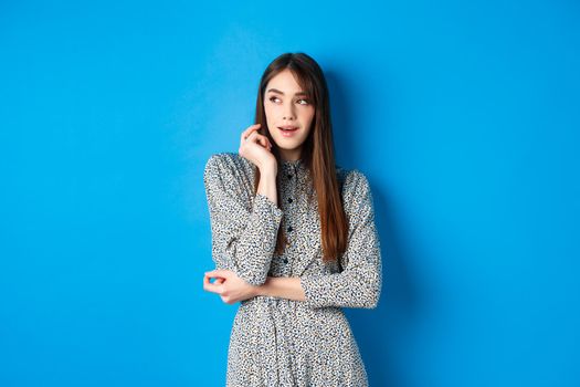 Pensive caucasian girl with long natural hair, wearing vintage dress, looking left with thoughtful face, standing on blue background.