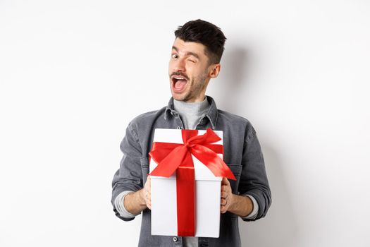 Funny young man holding valentines gift and winking at camera, making romantic surprise to lover, standing on white background.