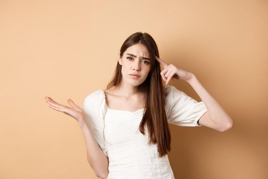 Are you stupid. Annoyed woman mocking person, pointing finger at head and shrugging confused, irritated by crazy ideas, standing on beige background.
