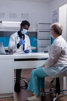 Medic of african ethnicity taking notes on papers for consultation with elder patient in medical cabinet. Black doctor consulting old woman at clinic during coronavirus pandemic