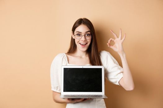 E-commerce. Excited young woman in glasses showing okay sign and laptop screen, recommending internet promo, standing on beige background.
