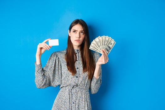 Shopping. Thoughtful girl looking up and biting lip, counting in mind or thinking, holding plastic credit card with dollar bills, standing on blue background.