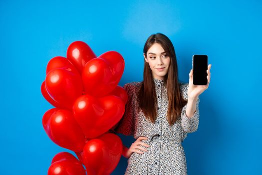 Valentines day. Pretty smiling woman in dress showing empty smartphone screen, standing near romantic balloons, blue background.