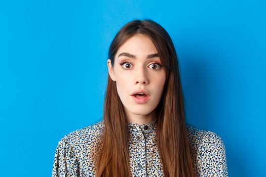 Close-up of excited pretty woman with long natural hair and makeup, drop jaw and gasping surprised, checking out awesome promo, hear amazing news, blue background.