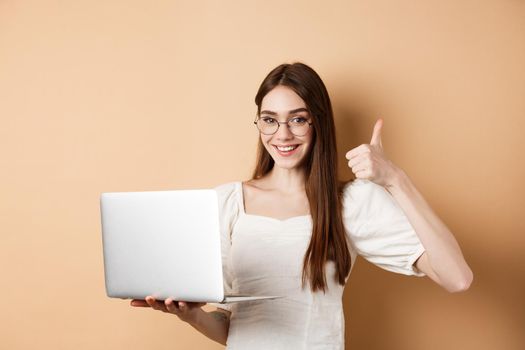 Smiling woman in glasses using laptop and showing thumb up, working on computer, standing on beige background.