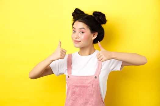 Beautiful asian girl with bright pink makeup, showing thumb up in approval and smiling satisfied, recommending promo, yellow background.