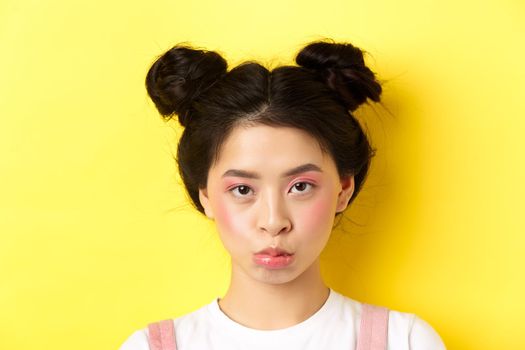 Close up portrait of moody asian girl pouting and looking upset at camera, standing wiht glamour makeup and hairbuns on yellow background.