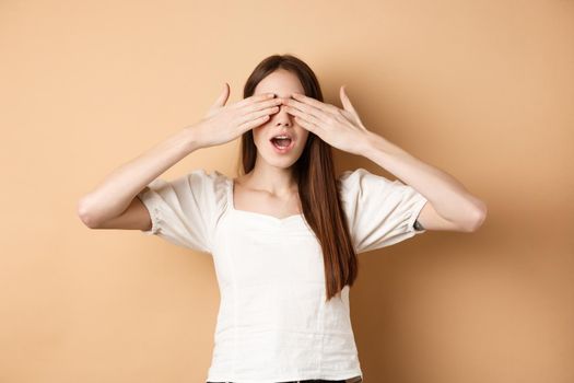 Happy romantic girl covering eyes and waiting for surprise, smiling while anticipating gift, standing on beige background.