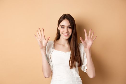 Attractive young woman show fingers number ten, smiling and looking confident, standing on beige background.
