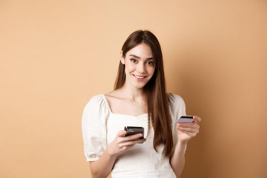 Smiling caucasian woman paying for online order, holding plastic credit card and mobile phone, looking carefree at camera, beige background.