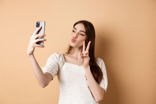 Fashion female blogger pucker lips and show v-sign at smartphone camera, taking selfie for social media, standing on beige background.