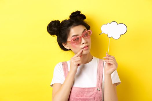 Summer and fashion concept. Stylish glamour asian girl in sunglasses, holding comment cloud party mask and looking aside, yellow background.