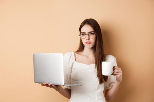 Tired working girl looking at laptop bored, drinking coffee while using computer for work, standing on beige background.