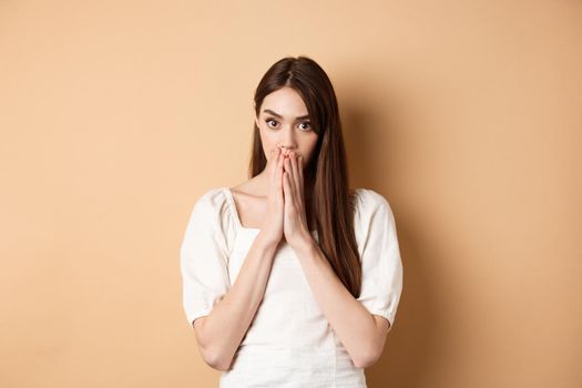 Omg shocking. Speechless woman covering mouth with hands and staring shocked at camera, gossiping, hear stunning news, beige background.