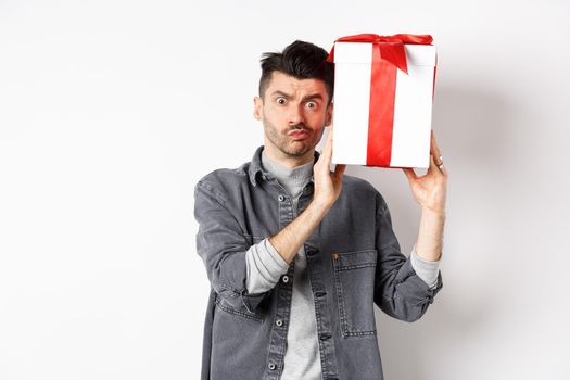 Intrigued young man looking confused at camera, trying to guess what inside gift box from lover, shaking present and stare puzzled, standing on white background. Valentines day concept.