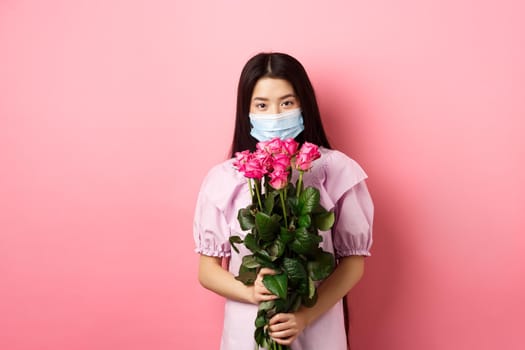 Young asian girl in medical mask holding flowers on Valentines day, receive bouquet of roses from lover, standing on pink background. Social distancing and covid-19 concept.