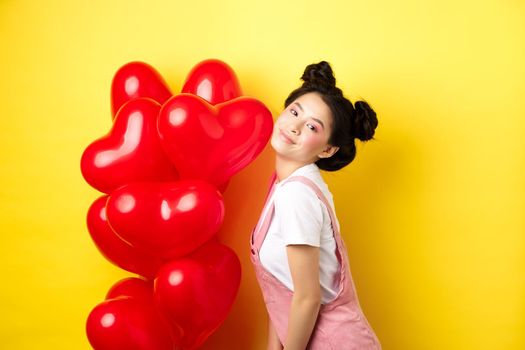 Valentines day concept. Stylish teenage asian girl posing near red hearts balloons, wear outfit for romantic date, standing happy on yellow background.