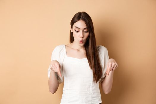 Wow look. Amazed beautiful girl checking out promo deal, pointing fingers down and gasping astonished, showing advertisement, beige background.