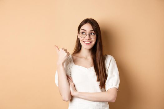 Happy woman in glasses smiling and pointing left, showing logo on empty space, standing on beige background.