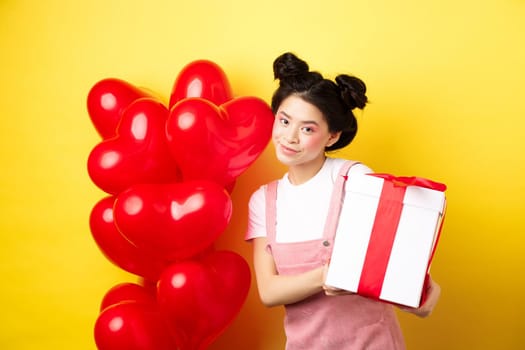 Happy Valentines day. Beautiful and stylish asian woman posing near heart balloons with lovely box with gift from lover, standing over yellow background.