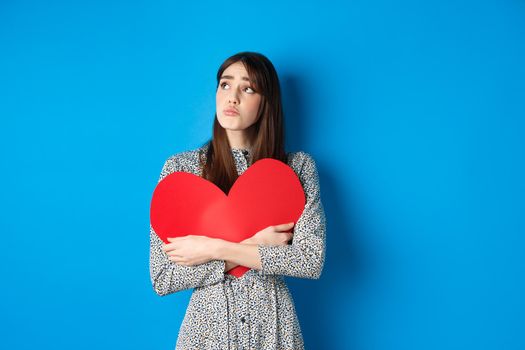 Valentines day. Sad heartbroken girl sobbing, looking aside at empty space and crying upset, hugging big red heart, feeling lonely without lover, standing alone on blue background.
