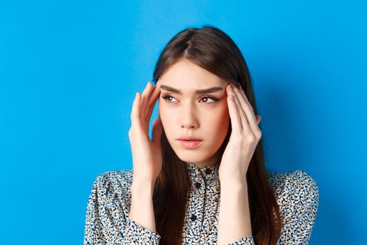 Close-up portrait of young woman feel sick, touching head temples and frowning from headache, having migraine, looking aside at logo, blue background.