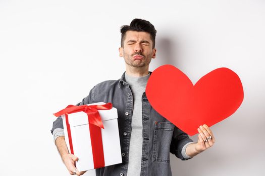 Sad and heartbroken man being rejected, crying and holding red heart with gift box, breakup on Valentines day, white background. Love and relationship concept.