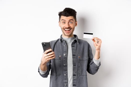 Online shopping concept. Excited man making purchase in internet, easy buying with plastic credit card and smartphone, smiling happy at camera, white background.