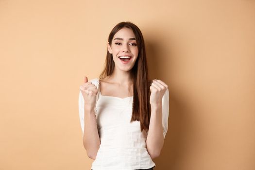 Happy smiling girl celebrating victory, making fist pump and say yes, achieve goal, triumphing or getting motivated, standing on beige background.