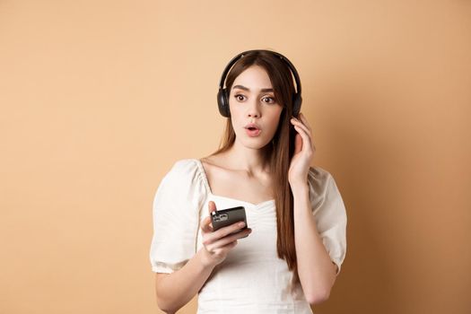 Wow awesome tunes. Excited young girl amazed with new song, listening music in headphones and look impressed, holding cell phone, beige background.